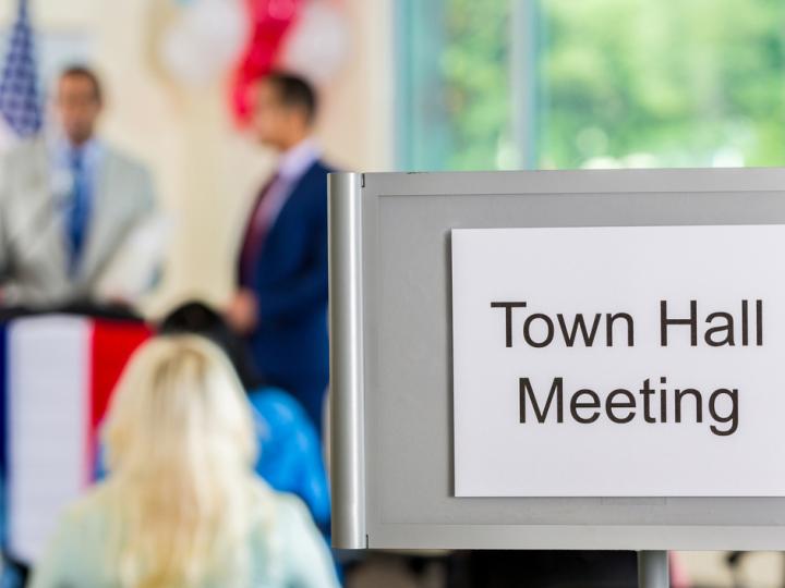 Image of a town hall meeting. In the foreground, a sign reads, &quot;Town Hall Meeting.&quot; The background is blurred, but figures are visible at a podium and in the audience.