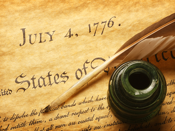 Image of the United States Declaration of Independence zoomed around the date July 4, 1776. A quill pen and ink pot are placed upon the document.