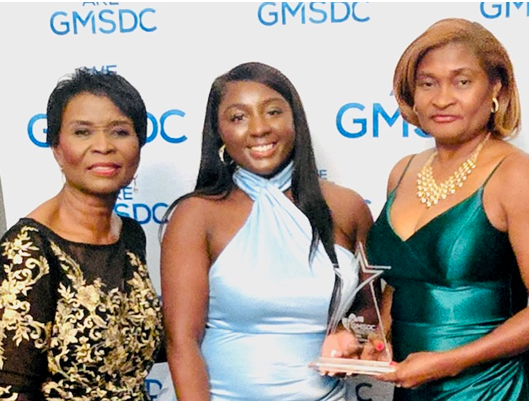 Three women stand in front of camera in evening gowns posing with award