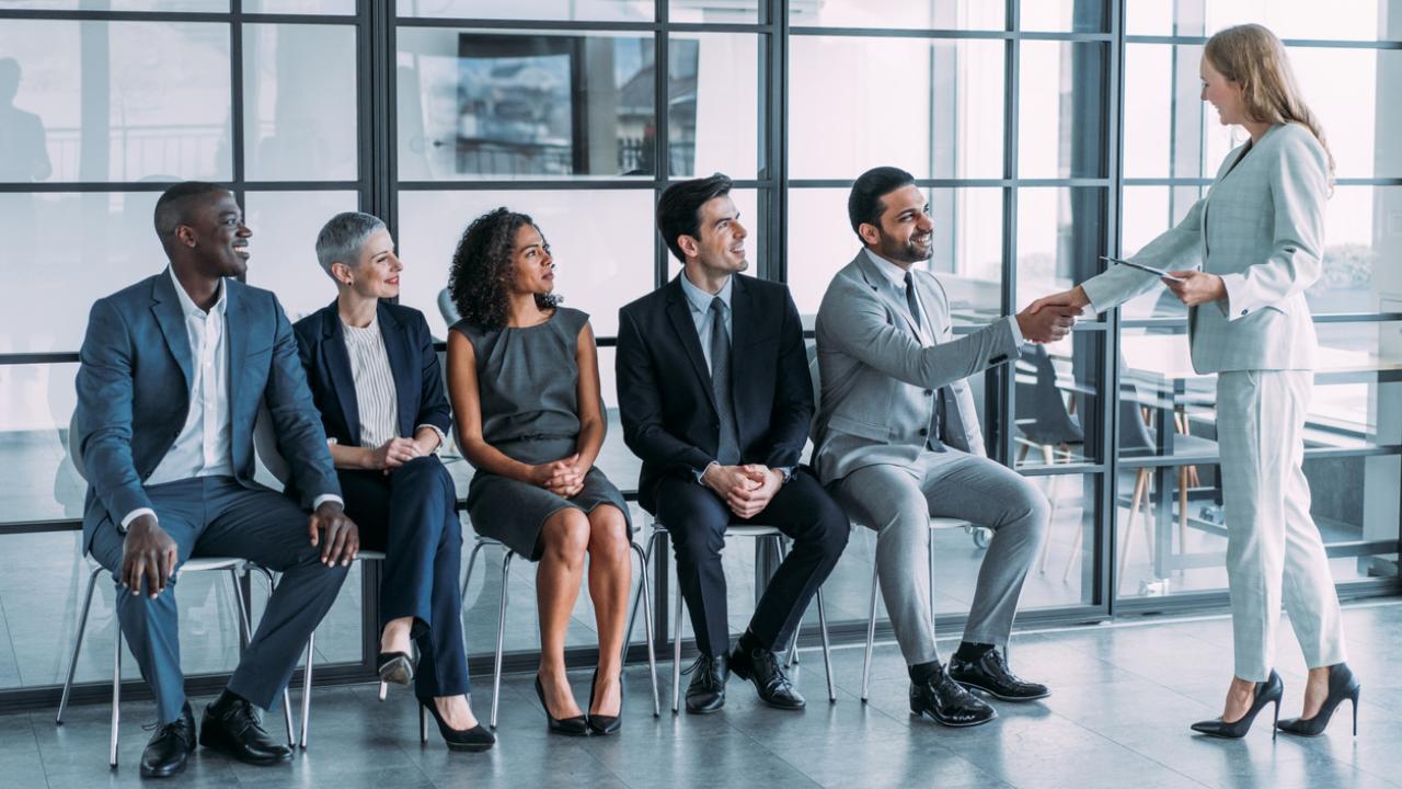 A row of five seated job candidates look up as a recruiter walks into the room.