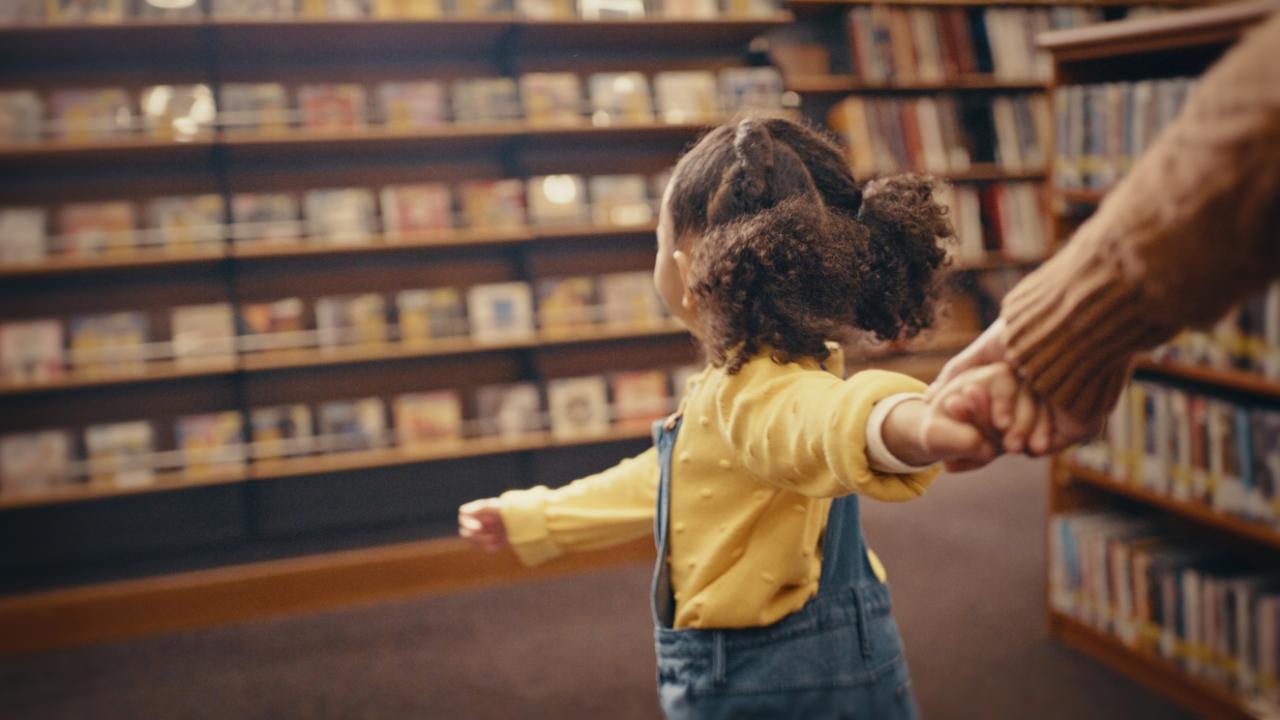 A young girl pulls an adult toward a set of books in a library.