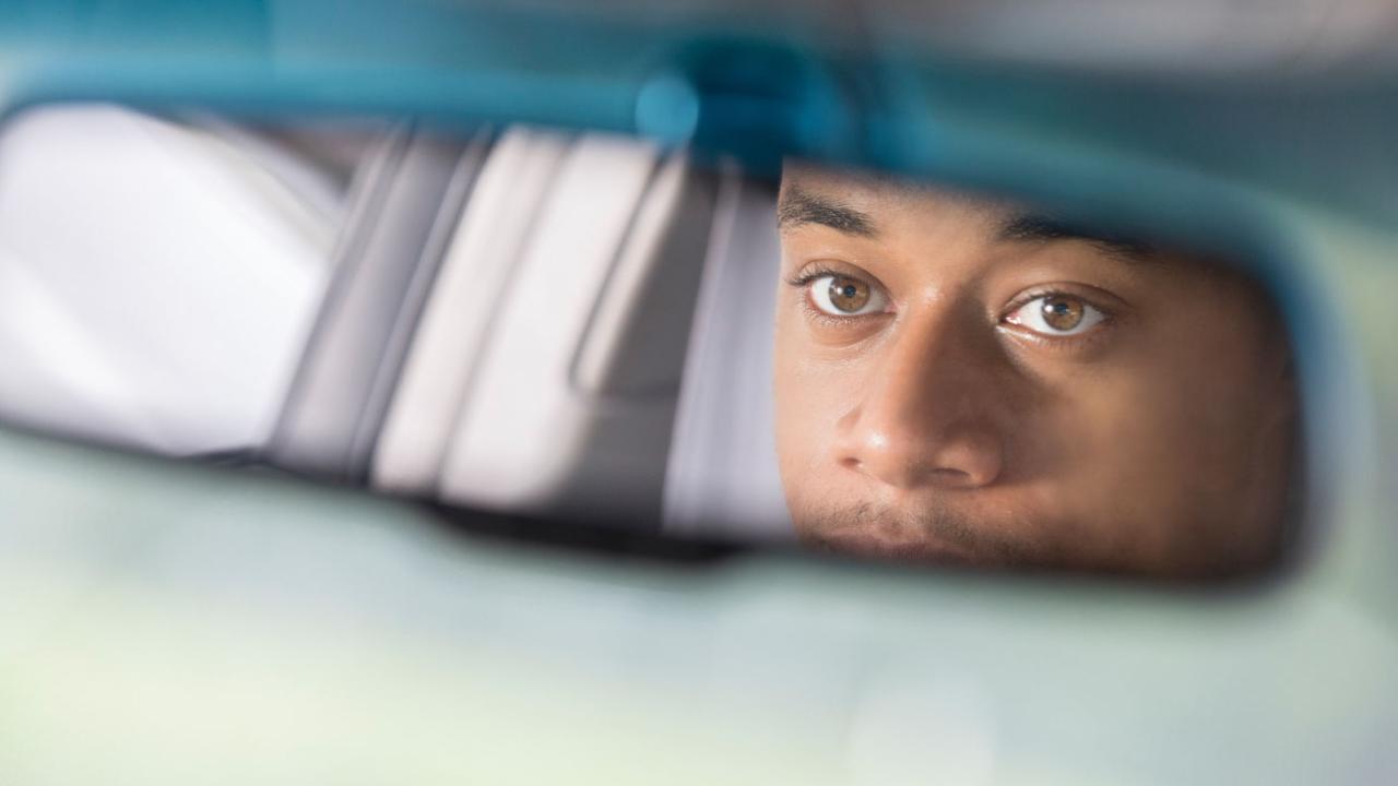 Image of a car&#039;s rear-view mirror, with a man looking into it. 