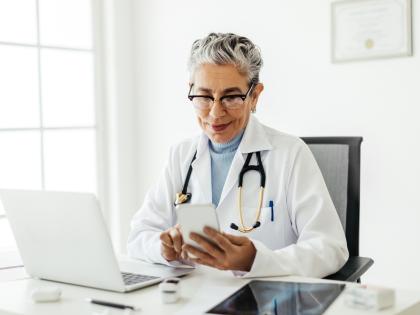 Image of a doctor looking something up on her phone while seated at a desk with a laptop computer.