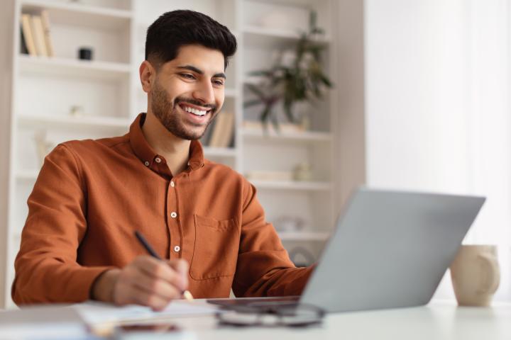 Man sitting at desk in front of laptop smiling and taking notes