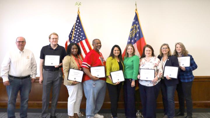 A group of several people, each holding a copy of their DOAS Leadership Award, stands before the United States and Georgia flags.