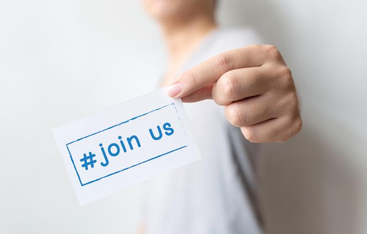 Foreground: A hand holds a card with the words &quot;join us.&quot; Background: A person, faded and mostly cropped out of frame.