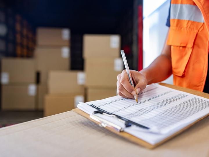 A person checks off items on a clipboard. Boxes are stacked in the background.