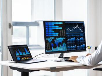 A desk with laptop and desktop computer, both showing charts, graphs, and other technical imagery.