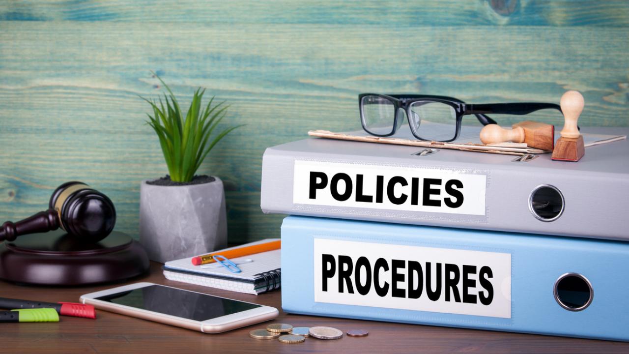 Image of a desk with items including two binders labeled "POLICIES" and "PROCEDURES."