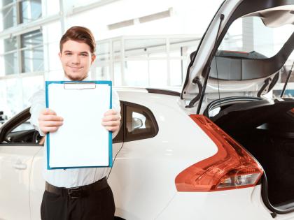 A smiling man holds out a clipboard. He is standing in front of a new sedan, its hatchback open.